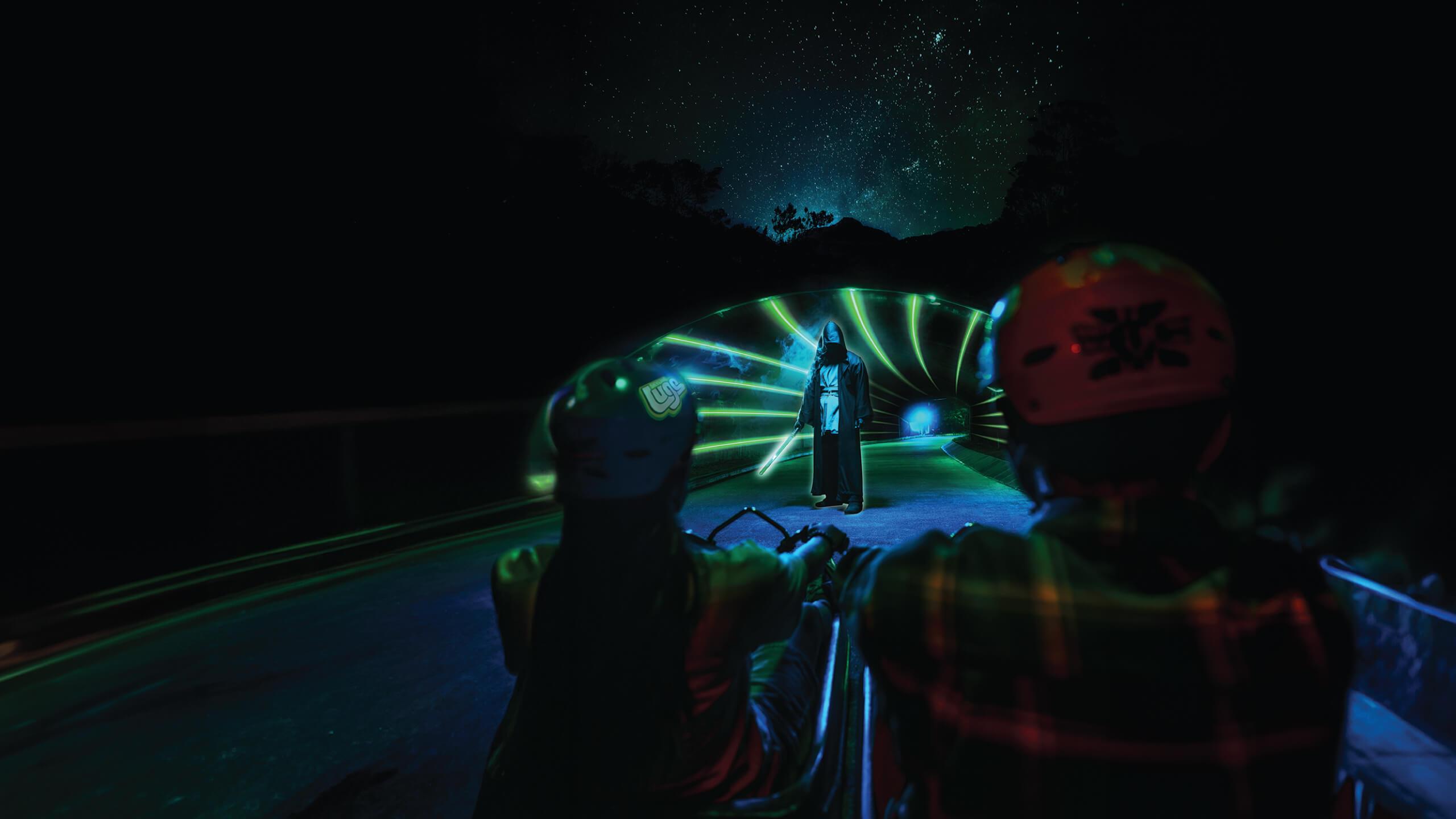 Two people Luge through an immersive light tunnel seeing a person holding a lightsabre.