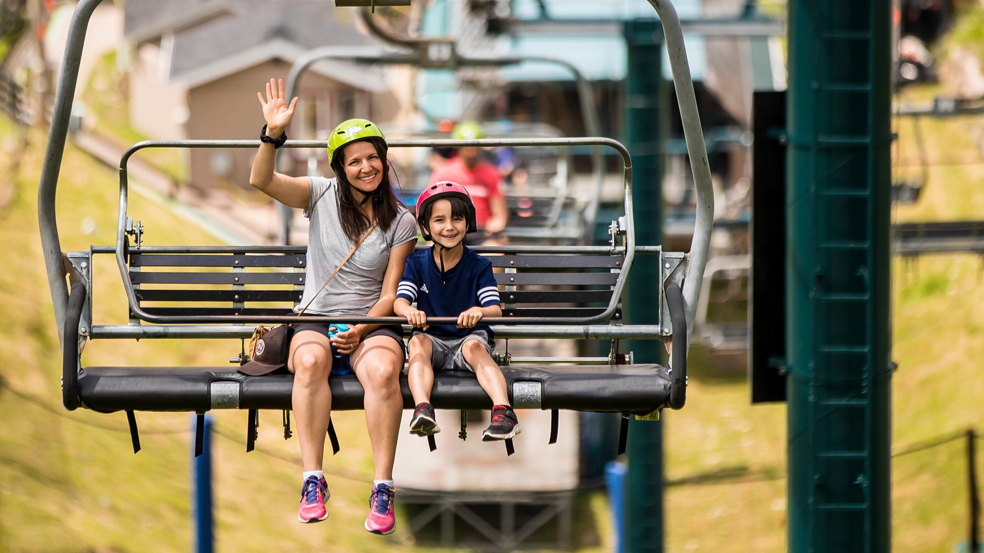 A mother and son smile and wave as they ride the chairlift to the top of the tracks.